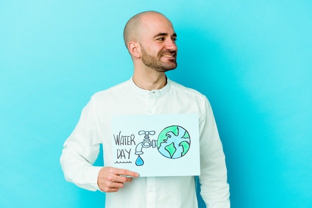 El hombre calvo caucásico joven que celebra el día mundial del agua aislado en azul mira a un lado sonriente, alegre y agradable.