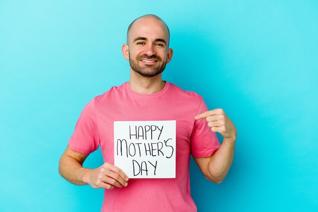 Hombre calvo caucásico joven que sostiene un cartel del día de madres feliz aislado en la persona de la pared azul que señala con la mano a un espacio de la copia de la camisa, orgulloso y confiado