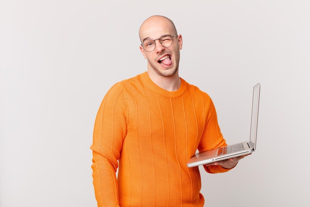 Hombre calvo con computadora con actitud alegre, despreocupada, rebelde, bromeando y sacando la lengua, divirtiéndose