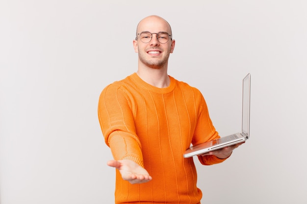 Hombre calvo con computadora sonriendo felizmente con mirada amigable, segura y positiva, ofreciendo y mostrando un objeto o concepto