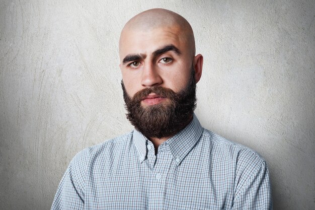 Un hombre calvo confiado con gruesas cejas negras y barba con camisa a cuadros con expresión sombría