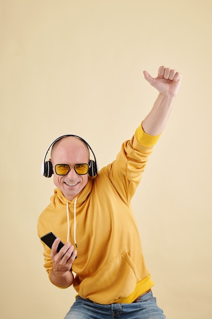 Hombre calvo feliz en auriculares y sudadera con capucha bailando con la música en su teléfono inteligente