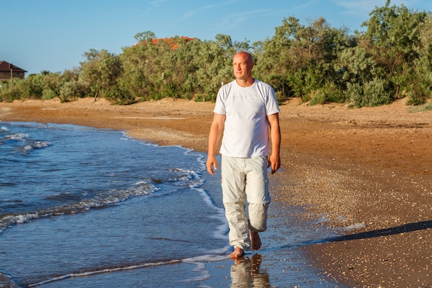 Hombre calvo feliz caminando en la playa en ropa blanca