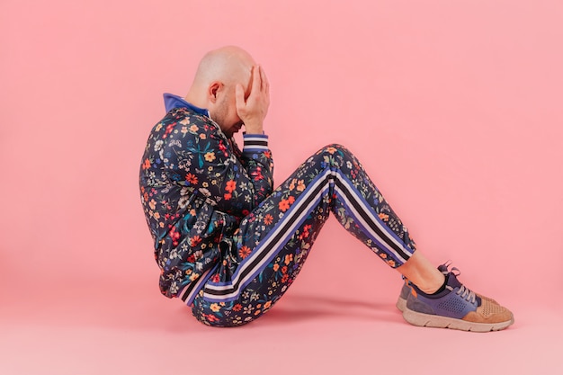 Hombre calvo en ropa de moda sentado con las manos en su mirada sobre fondo rosa.