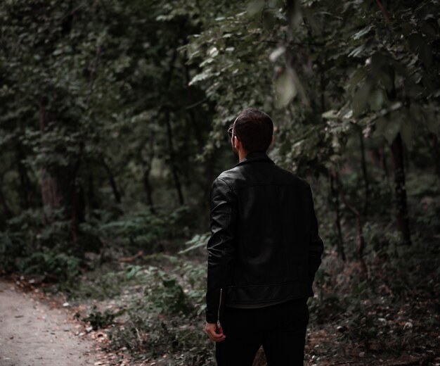Un hombre camina por el bosque con una chaqueta de cuero y gafas de sol Vista posterior