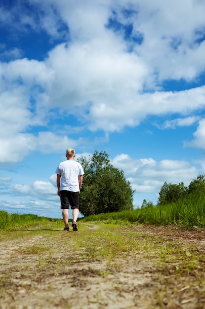 El hombre camina por un sendero cerca de un bosque en pantalones cortos oscuros y una camiseta blanca.