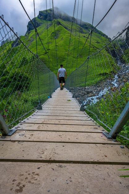 Un hombre camina sobre un puente colgante entre montañas cubiertas de hierba