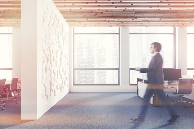 Foto hombre caminando en el interior de una oficina moderna con paredes blancas hexagonales, suelo enmoquetado, mesas de ordenador blancas y de madera y sala de reuniones a la izquierda. desenfoque de imagen tonificada