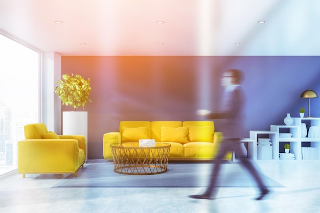 Hombre caminando en el interior de un salón moderno con paredes grises, suelo blanco, sillón amarillo y sofá parado cerca de una mesa baja redonda y estanterías blancas. Imagen tonificada borrosa