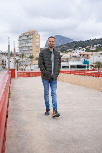 Hombre caminando por un paseo con las manos en los bolsillos de la chaqueta. Sonríe mientras camina. En un día nublado. Lleva una chaqueta verde, jeans y un suéter gris. Alicante, javea