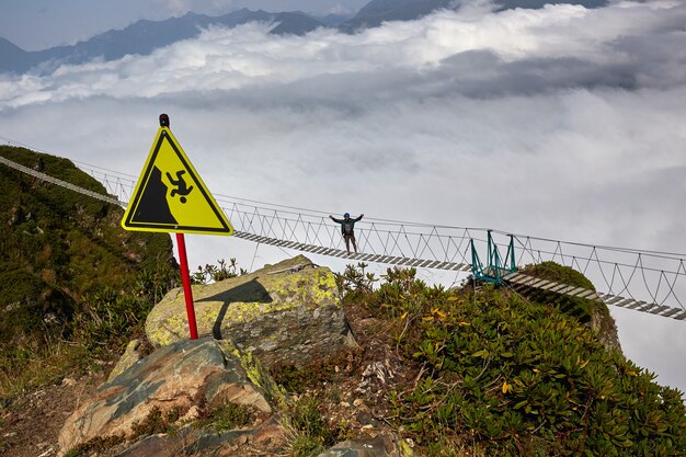 Foto hombre caminando sobre el puente colgante y mirando las montañas nubladas a continuación.