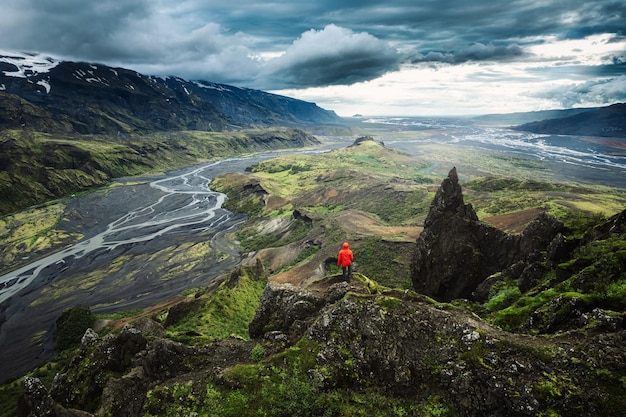 Hombre caminante con chaqueta roja parado en la cima de Valahnukur rodeado por una montaña volcánica y el río Krossa en las Tierras Altas de Islandia en Thorsmork