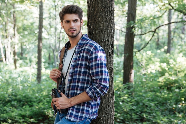 Hombre en camisa con binoculares y mochila cerca del árbol.