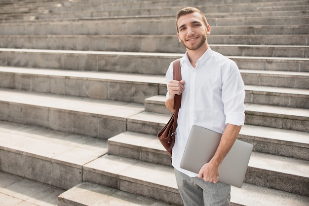 Foto hombre de camisa blanca sosteniendo una computadora portátil y sonriendo a la cámara