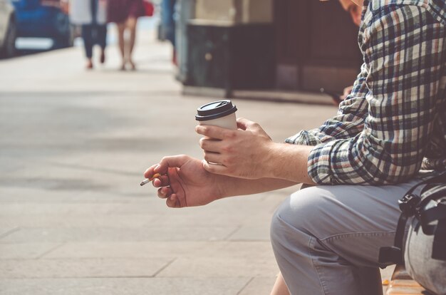 Foto el hombre con una camisa a cuadros está sentado en la calle y sostiene una taza de café de cartón y un cigarrillo. imagen tonificada con enfoque suave.