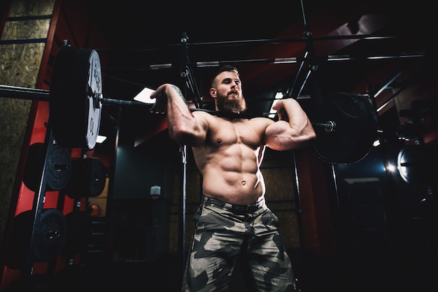 Hombre sin camisa culturista poderosamente motivado sosteniendo una barra pesada