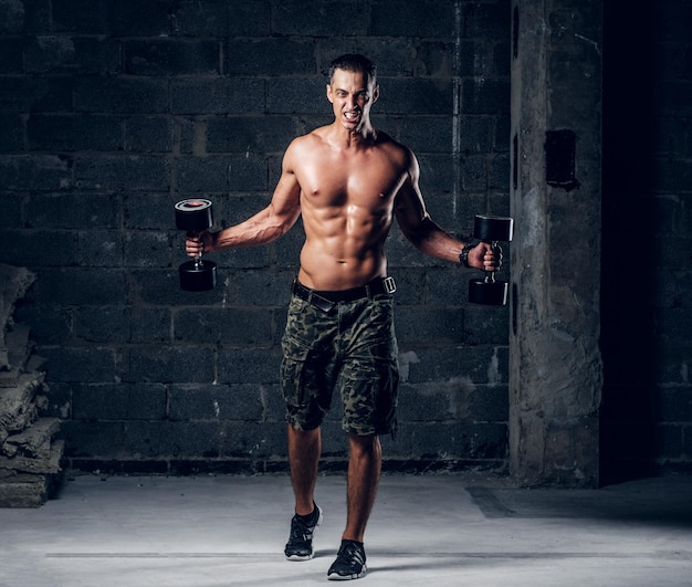 Un hombre sin camisa gritando con gorra está haciendo ejercicios con pesas en un estudio fotográfico oscuro.