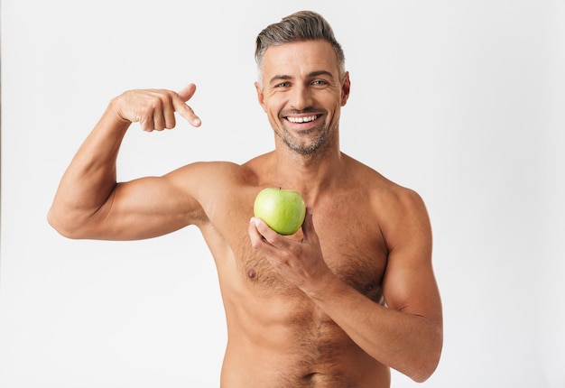 Hombre sin camisa guapo seguro de pie aislado en blanco, flexionando bíceps, mostrando manzana verde