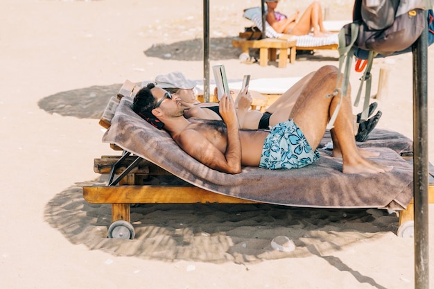 Hombre sin camisa leyendo un libro mientras está tendido en una silla de descanso en la playa