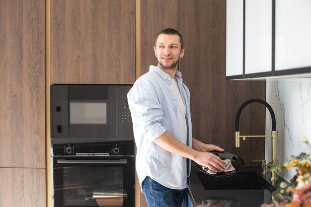 Un hombre con una camisa limpia los platos con una toalla de pie en la cocina Tareas domésticas