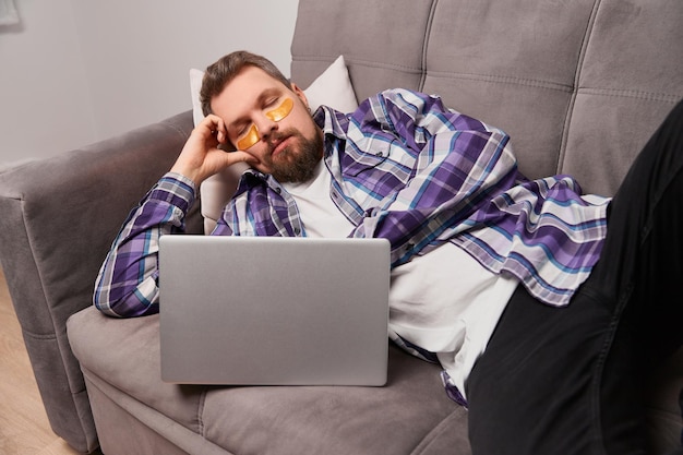 Hombre con camisa morada y parches en los ojos durmiendo en el sofá con una rutina de autocuidado portátil