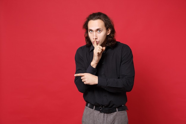 Hombre con camisa negra posando aislado en un retrato de estudio de fondo rojo. Concepto de estilo de vida de las emociones de las personas. El espacio de copia simulado susurra chismes dice secretos con un gesto de la mano, señalando con el dedo índice a un lado.