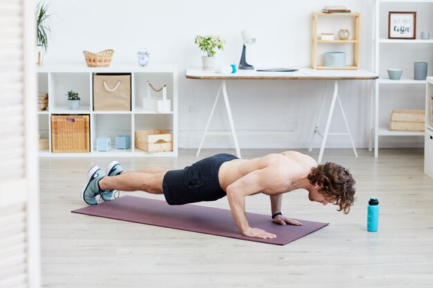 Hombre sin camisa sano haciendo flexiones en la colchoneta de ejercicios durante el entrenamiento deportivo en casa