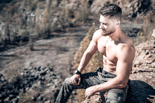 Hombre sin camisa sentado en una roca Hombre atleta o culturista en las montañas Paisaje natural Clima soleado