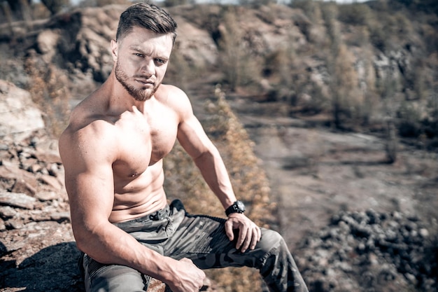 Hombre sin camisa sentado en una roca Hombre atleta o culturista en las montañas Paisaje natural Clima soleado