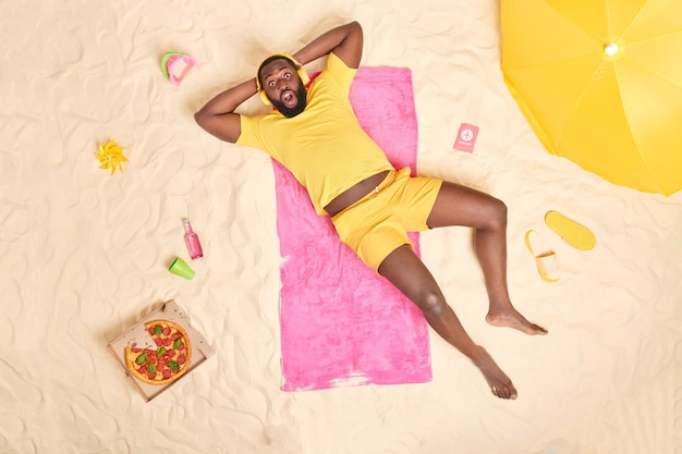 El hombre con camiseta amarilla y pantalones cortos se encuentra sobre una toalla rosa rodeada de accesorios de playa escucha música a través de auriculares