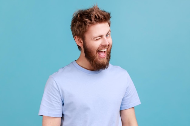 Hombre de camiseta azul de buen humor sonriendo y guiñando un ojo a la cámara mantiene la boca abierta coqueteando expresión