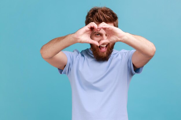 Hombre con camiseta azul mirando a través de las manos en forma de corazón mostrando un gesto romántico confesión de amor