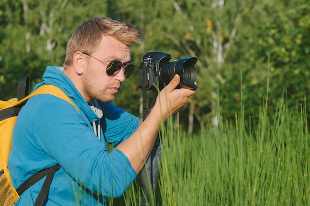 Un hombre con una camiseta azul y una mochila amarilla sostiene una cámara de video y fotografía profesional en sus manos. En el contexto de la naturaleza verde y el bosque.