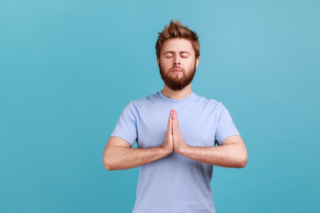 Hombre en camiseta azul de pie con los ojos cerrados y gesto de oración meditando concentrando pensamientos