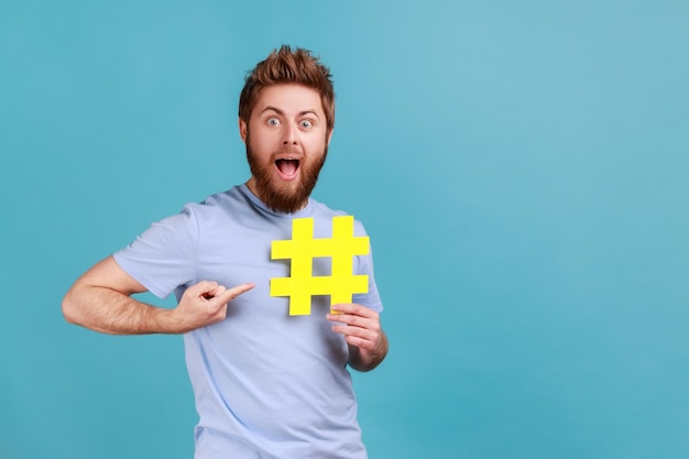 Hombre en camiseta azul señalando con el dedo el hashtag en la mano con la boca abierta y la búsqueda del sitio web de ojos grandes