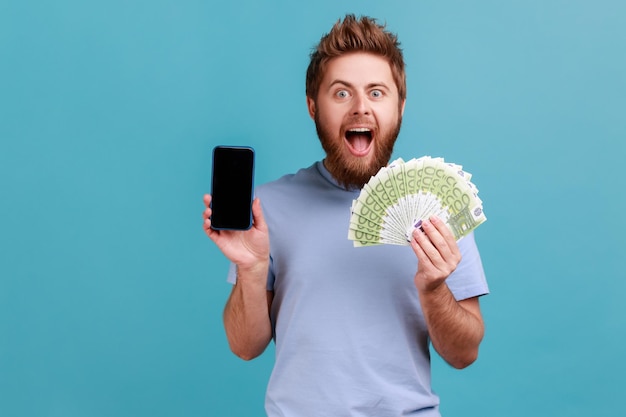 Hombre en camiseta azul sosteniendo billetes en euros y teléfono celular con pantalla vacía simulada para publicidad