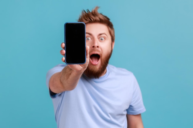 Hombre en camiseta azul sosteniendo el teléfono celular y mirando a la cámara con ojos grandes y boca abierta