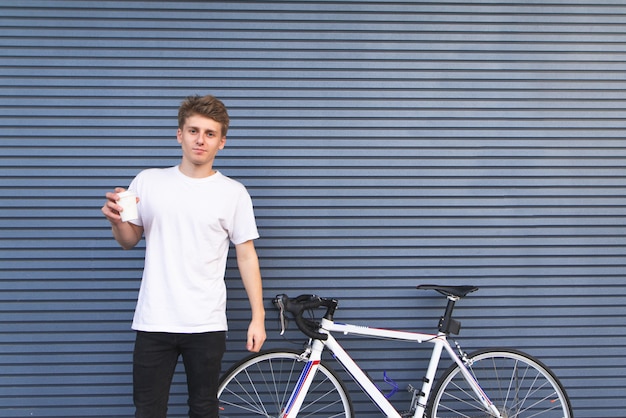 El hombre con una camiseta blanca está junto a una bicicleta con una taza de café en sus manos