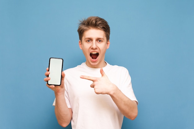 Un hombre con una camiseta blanca con un teléfono inteligente en la mano mira a la cámara con una cara feliz y señala con el dedo a la pantalla blanca.