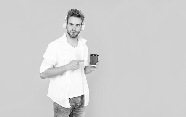 El hombre con camiseta blanca usa auriculares señalando con el dedo la taza de café con una bebida de fondo amarillo