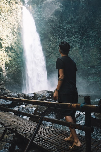 Foto hombre de camiseta negra parado en un puente de madera cerca de cascadas foto