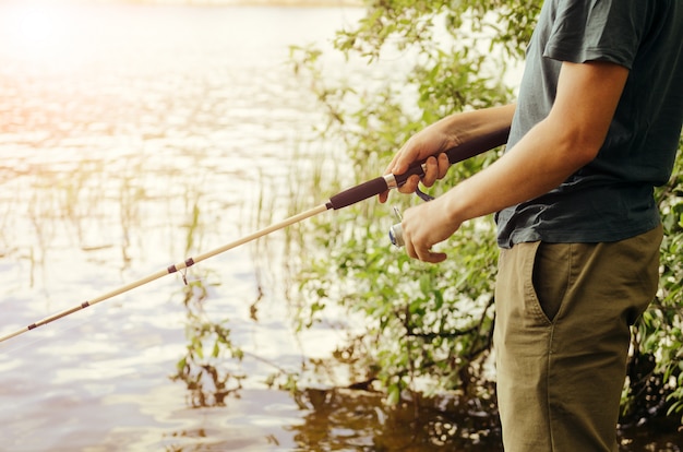 Un hombre con una camiseta y pantalones oscuros está pescando en el lago con una caña de pescar blanca contra el fondo de arbustos y hierba. Concepto de ocio activo.