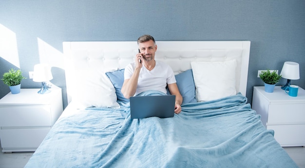 Foto hombre canoso hablando por teléfono y trabajando en la computadora portátil en la cama