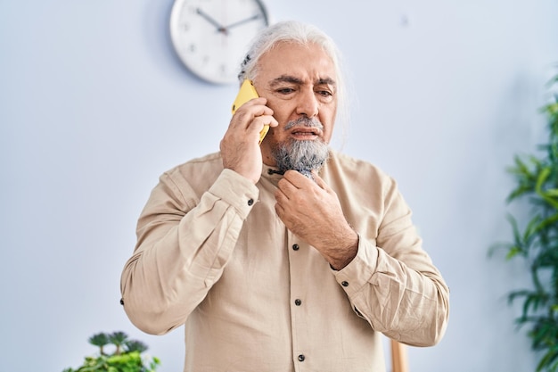 Hombre canoso de mediana edad hablando por teléfono inteligente con expresión preocupada en casa