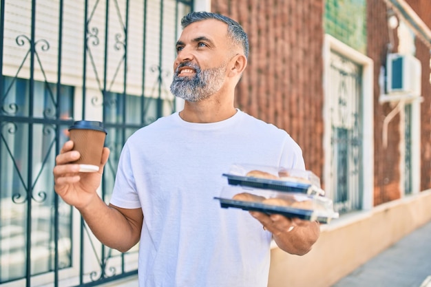 Hombre canoso de mediana edad sonriendo feliz sosteniendo una taza de café y comida para llevar en la ciudad