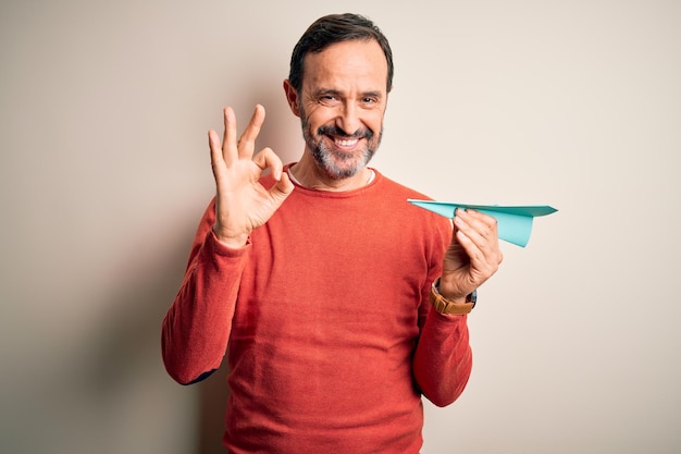 Hombre canoso de mediana edad sosteniendo aviones de papel de pie sobre fondo blanco aislado haciendo el signo de ok con los dedos símbolo excelente