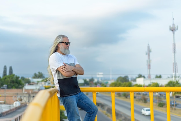 Hombre canoso de pelo largo con gafas oscuras en un puente peatonal Paisaje urbano en el fondo