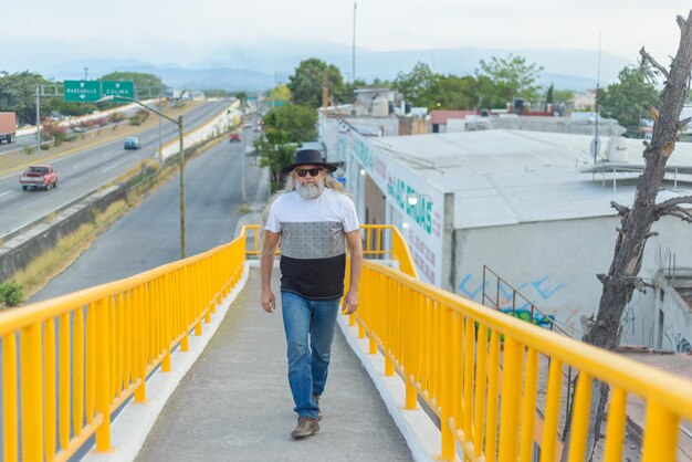 Hombre canoso de pelo largo con un sombrero negro en un puente peatonal con vistas a la ciudad