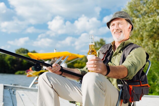 Hombre canoso radiante sonriendo mientras pesca y bebe cerveza