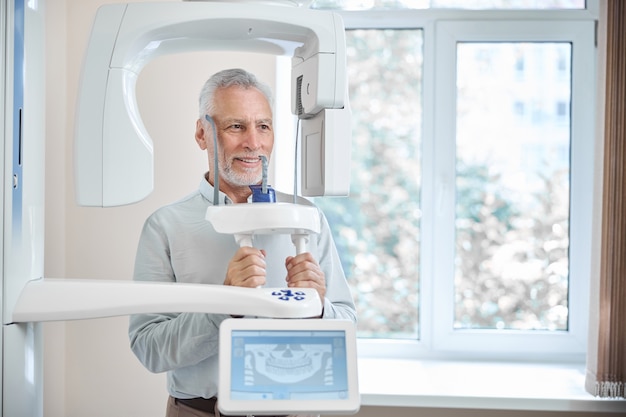 Hombre canoso sonriendo mientras está de pie junto a un radiógrafo panorámico en una clínica dental